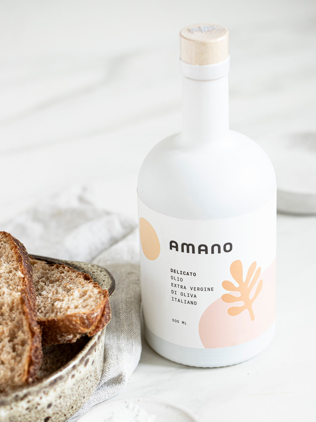 Amano Delicato Extra Vergine Olivenöl mit weißem Hintergrund, knusprigem Brot und Salzkörnern in einer Schale
